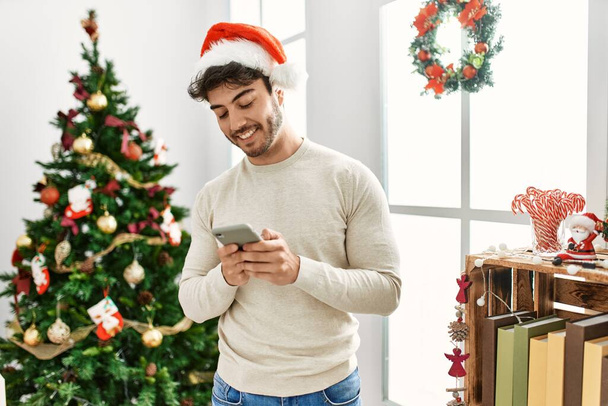Joven hombre hispano usando sombrero de navidad usando smartphone en casa. - Foto, imagen