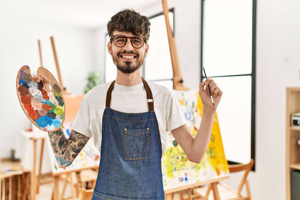 Jeune artiste hispanique homme souriant heureux tenant pinceau et palette au studio d'art. - Photo, image