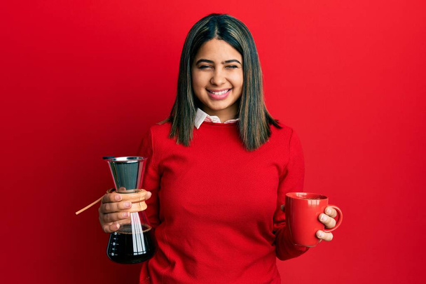 Молодая латиноамериканка с кофейным фильтром и чашкой подмигивания смотрит в камеру с сексуальным выражением лица, веселым и счастливым лицом.  - Фото, изображение