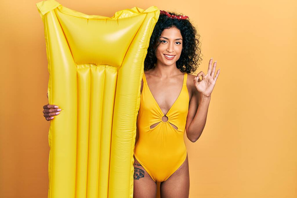 Young latin girl wearing swimsuit and holding summer mattress float doing ok sign with fingers, smiling friendly gesturing excellent symbol  - Photo, Image