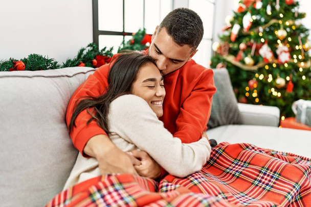 Jovem casal hispânico sorrindo feliz e abraçando sentado no sofá pela árvore de natal em casa. - Foto, Imagem