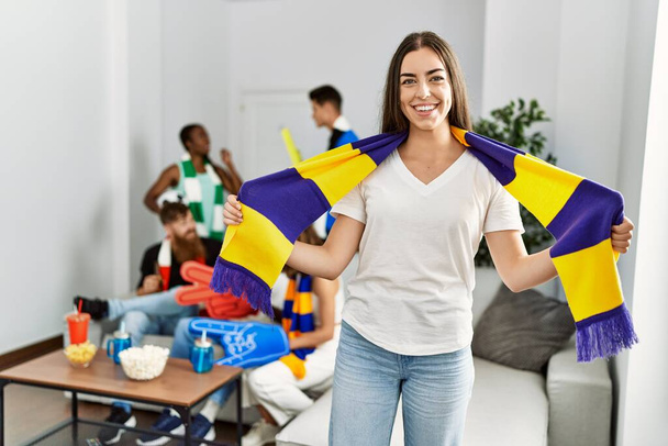 Joven futbolista mujer hooligan sonriendo feliz sosteniendo equipo bufanda en casa - Foto, Imagen