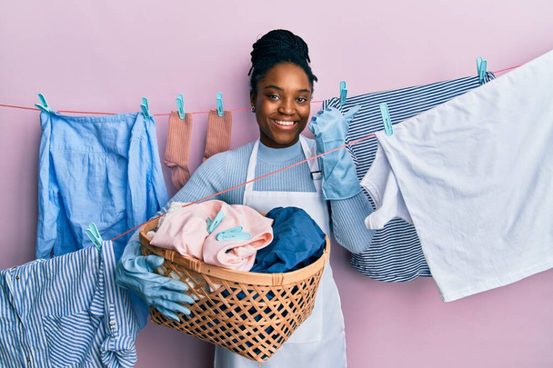 Afroamerikanerin mit geflochtenen Haaren beim Wäschewaschen, Weidenkorb haltend, Daumen zur Seite zeigend, glücklich lächelnd mit offenem Mund  - Foto, Bild