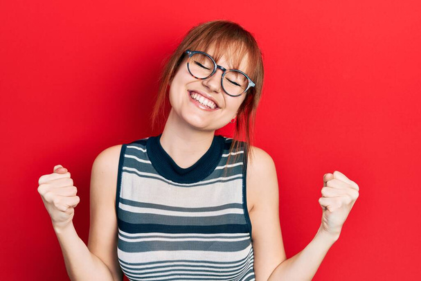 Die rothaarige junge Frau in lässiger Kleidung und Brille ist sehr glücklich und aufgeregt und macht Siegergeste mit erhobenen Armen, lächelt und schreit nach Erfolg. Festkonzept.  - Foto, Bild