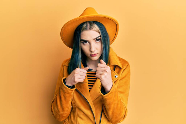 Jovem menina moderna vestindo chapéu amarelo e jaqueta de couro pronto para lutar com gesto de defesa punho, rosto irritado e chateado, com medo do problema  - Foto, Imagem