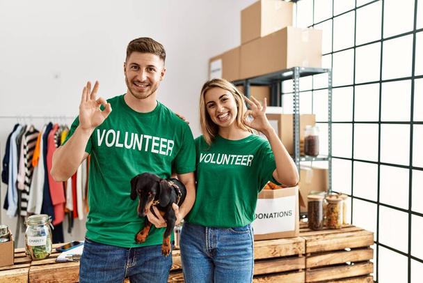 Jong stel met schattige hond dragen vrijwilliger t-shirt bij donaties staan doen ok teken met de vingers, glimlachend vriendelijk gebaar uitstekend symbool  - Foto, afbeelding