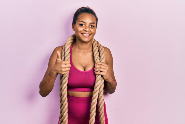 Fiatal afro-amerikai lány edzés harci kötél mosolygós boldog és pozitív, hüvelykujj fel csinál kiváló és jóváhagyási jel  - Fotó, kép