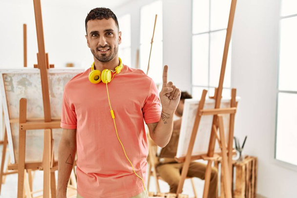 Young hispanic man at art studio showing and pointing up with finger number one while smiling confident and happy.  - Photo, Image
