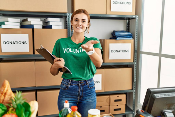 Młoda brunetka kobieta nosząca koszulkę wolontariusza na darowiznach stoją uśmiechnięte radosne podając dłoń podając pomoc i akceptację.  - Zdjęcie, obraz
