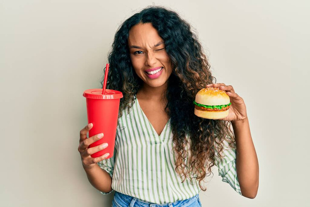 Mujer latina joven comiendo una sabrosa hamburguesa clásica y gaseosa guiñando el ojo a la cámara con expresión sexy, cara alegre y feliz.  - Foto, Imagen