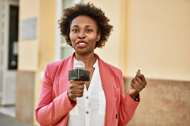 Joven mujer afroamericana periodista sosteniendo micrófono de reportero hablando y sonriendo a la cámara para noticias de televisión - Foto, Imagen