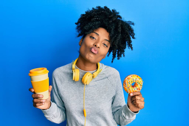 Αφροαμερικανή γυναίκα με αφρο-μαλλιά να τρώει ντόνατ και να πίνει καφέ κοιτάζοντας την κάμερα να φυσάει ένα φιλί να είναι όμορφη και σέξι. έκφραση αγάπης.  - Φωτογραφία, εικόνα