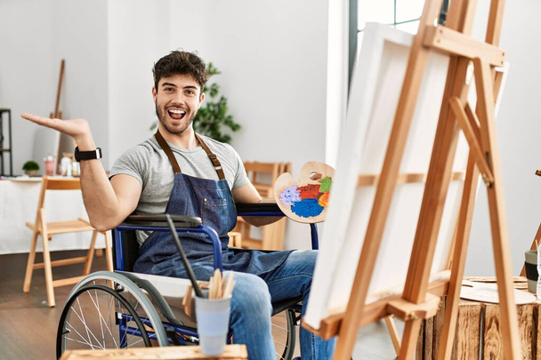 Νεαρός Ισπανόφωνος κάθεται σε αναπηρική καρέκλα ζωγραφική στο στούντιο τέχνης δείχνοντας στην άκρη με τα χέρια ανοιχτά παλάμες δείχνει αντίγραφο χώρο, παρουσιάζοντας διαφήμιση χαμογελώντας ενθουσιασμένος χαρούμενος  - Φωτογραφία, εικόνα