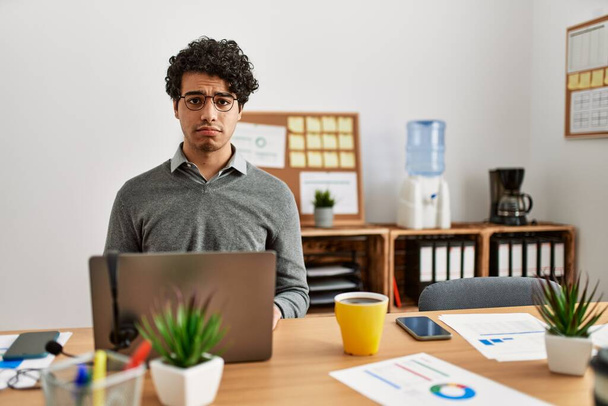 Ein junger hispanischer Mann im Business-Stil sitzt deprimiert auf dem Schreibtisch im Büro und sorgt sich um sein Leben, weint wütend und ängstlich. Traurige Miene.  - Foto, Bild