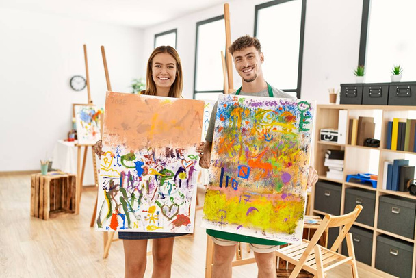 Young hispanic artist couple smiling happy showing canvas draw at art studio. - Foto, afbeelding