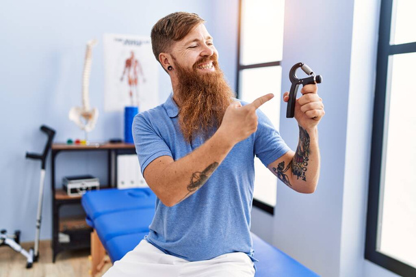 Young redhead man wearing physiotherapist uniform holding hand grip at physiotherapy clinic - Photo, Image