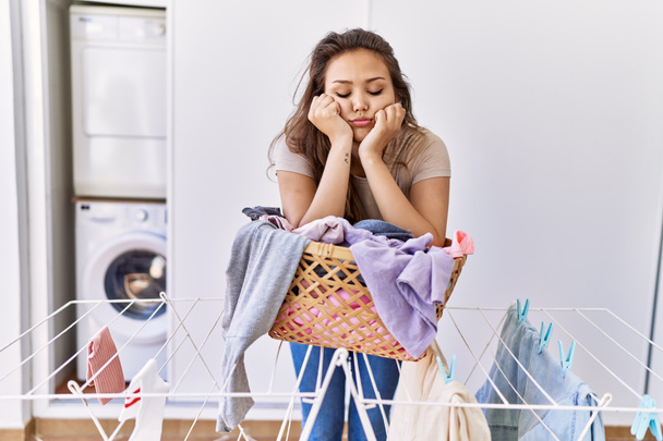 Young hispanic girl tired leaning on laundy basket at home - 写真・画像