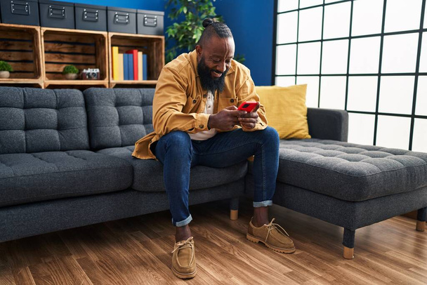 młody Afroamerykanin człowiek za pomocą smartfona siedzi na kanapie w domu - Zdjęcie, obraz