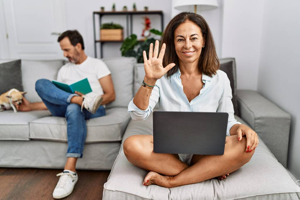 İspanyol orta yaşlı çift evde, dizüstü bilgisayar kullanan kadın beş numarayı gösterip işaret ederken kendinden emin ve mutlu bir şekilde gülümsüyor..  - Fotoğraf, Görsel