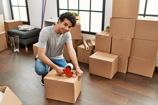 Νεαρός Ισπανός χαμογελάει ευτυχισμένος συσκευάζοντας κουτί στο νέο σπίτι. - Φωτογραφία, εικόνα