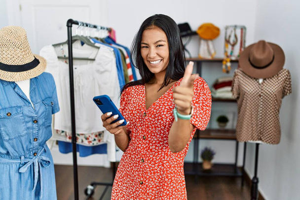 Молодая латиноамериканка работает в розничном бутике, используя смартфон, указывая пальцем на камеру со счастливым и смешным лицом. хорошая энергия и вибрации.  - Фото, изображение