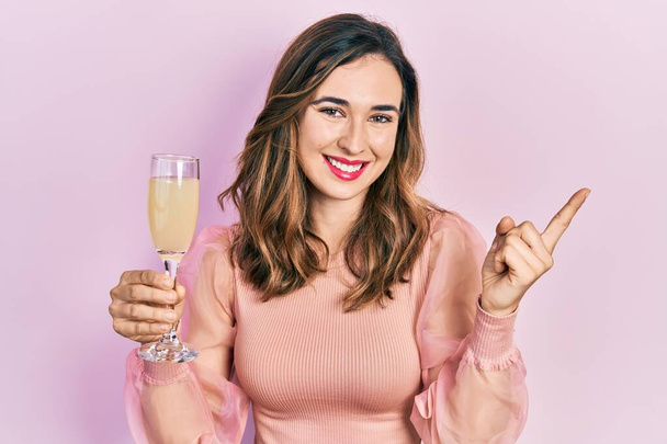 Giovane ragazza ispanica bere un bicchiere di champagne frizzante sorridente felice indicando con mano e dito di lato  - Foto, immagini