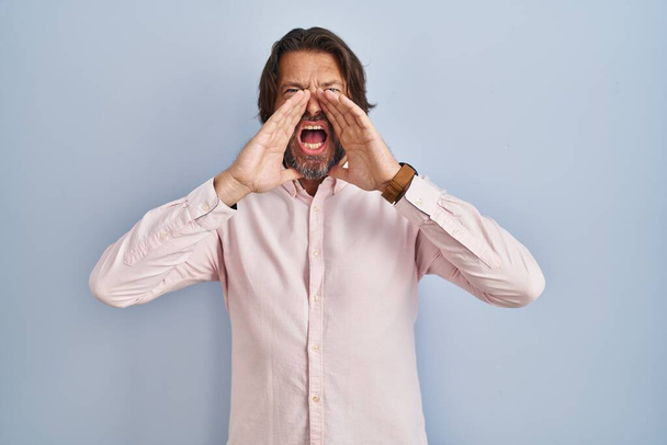 Όμορφος μεσήλικας άντρας που φοράει κομψό φόντο πουκάμισο φωνάζοντας θυμωμένος δυνατά με τα χέρια πάνω από το στόμα  - Φωτογραφία, εικόνα