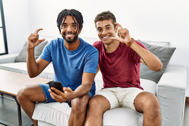 Молодые латиноамериканцы используют смартфон, сидя дома на диване, улыбаясь и уверенно жестикулируя рукой, делая знак малого размера с пальцами и камерой. концепция измерения.  - Фото, изображение