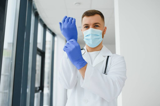 Young smiling Caucasian male doctor in white uniform, with eyeglasses and stethoscope around neck putting rubber gloves before patient examination. - Fotografie, Obrázek