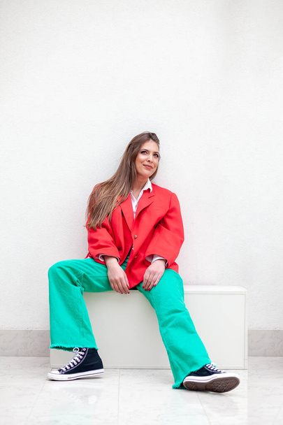 young woman sitting on white box wearing red jacket and green trousers - Foto, afbeelding