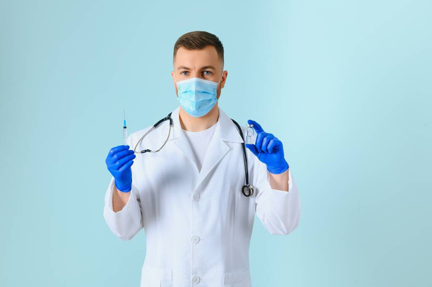 Koronavirüse karşı aşı. COVID-19 aşıları test ediliyor. Tıbbi maskeli bir erkek doktor mavi arka planda COVID-19 aşısı olan bir şırınga dolduruyor. Doktorun elleri şırıngayı yakın çekimde tutuyor.. - Fotoğraf, Görsel
