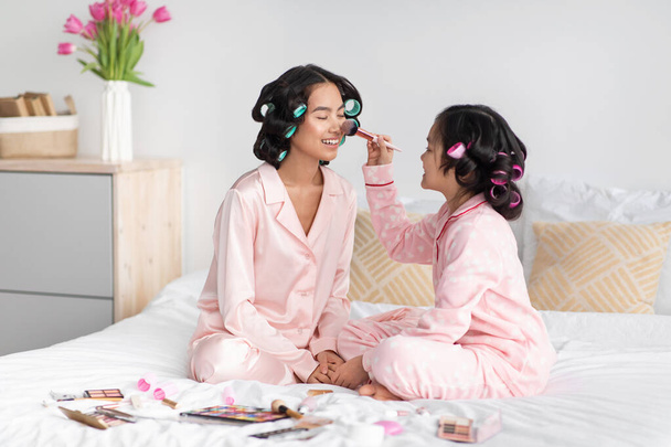 Fröhliches chinesisches Teenager-Mädchen mit Lockenwicklern im Haar appliziert Kosmetik auf das Gesicht der Millennial-Frau im rosa Pyjama - Foto, Bild