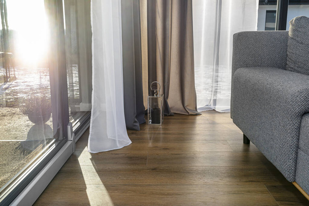 Moderno y sencillo salón en la casa con grandes ventanales y cortinas dobles, un sofá gris visible y una pequeña linterna. - Foto, Imagen