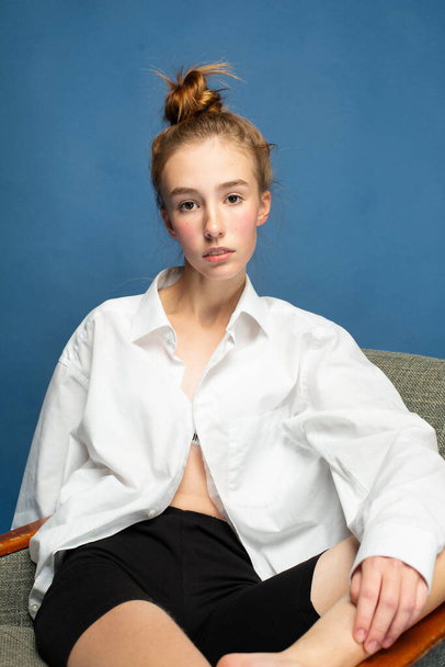Sitting girl with red hair and white skin isolated on blue in shirt - Foto, afbeelding