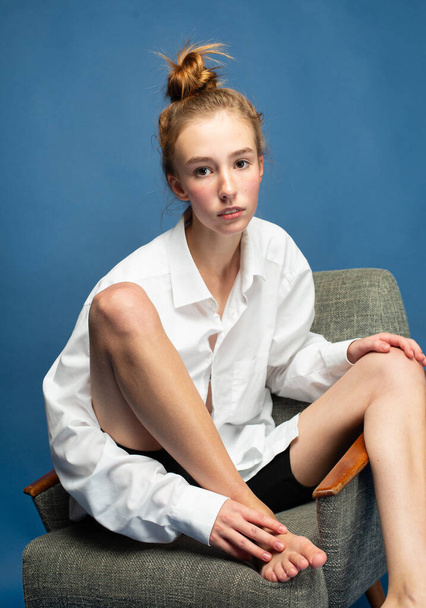 Tender girl with red hair and white skin isolated on blue sitting on chair - Foto, afbeelding