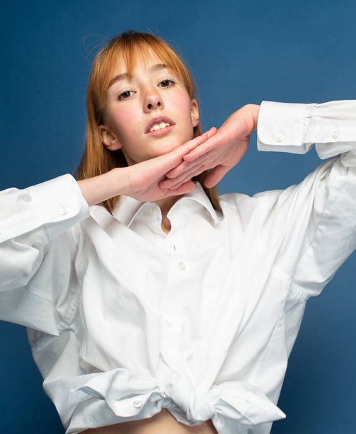 Girl with red hair and white skin isolated on blue with hands under her chin - Foto, afbeelding