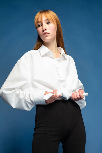 Standing girl with red hair and white skin isolated on blue in white tied shirt and black trousers - Foto, Bild