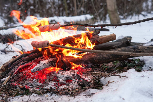 Embutidos en el palo a la parrilla en el fuego. Bosque de invierno. - Foto, imagen