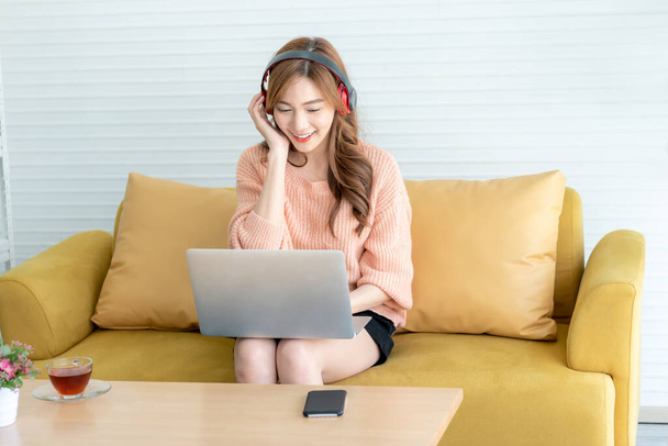 Asian woman, attractive and young Sitting on the sofa, wearing earphone and  work from home Which use notebook computers, to work from home and business online concept - Photo, Image