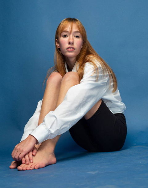 Sitting on the floor girl with red hair and white skin isolated on blue in white shirt and black breeches - Fotó, kép