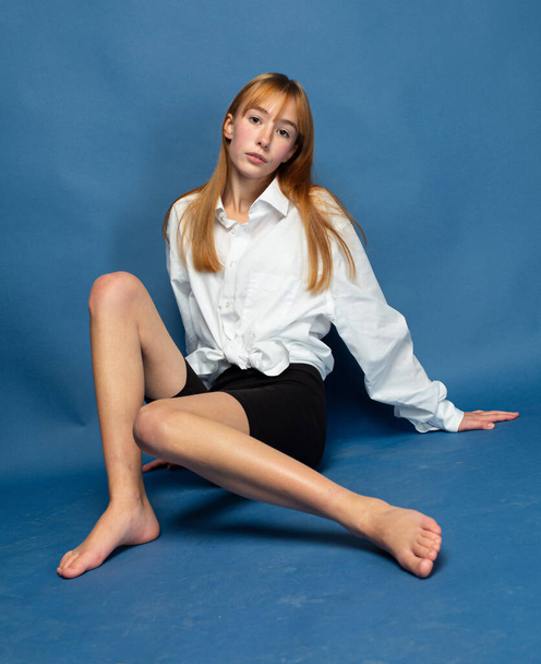 Sitting on the floor girl with red hair and white skin isolated on blue in white shirt and black breeches - Photo, Image