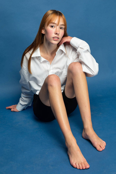 Slim sitting on the floor girl with red hair and white skin isolated on blue in white shirt - Fotó, kép