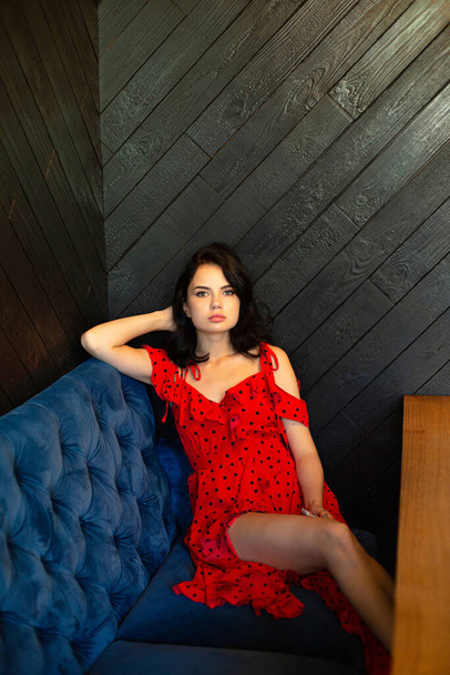 Young dark haired woman with white skin in dark wooden room sitting on a sofa with hand behind her head - Foto, Imagen
