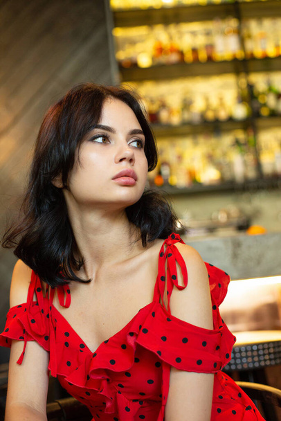 Portrait of young dark haired woman with white skin in bar in red dress - Photo, image