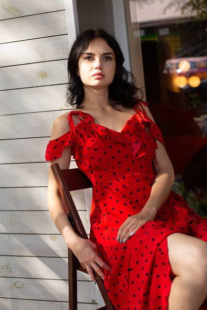 Sitting on chair young dark haired woman with white skin in red dress near cafe outside - Foto, afbeelding