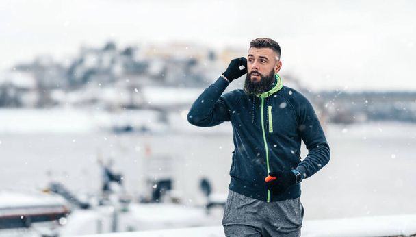 Handsome middle age man with a beard using his bluetooth era buds before start running and exercising outside on extremely cold and snowy day. Sport and fitness motivation theme. - 写真・画像