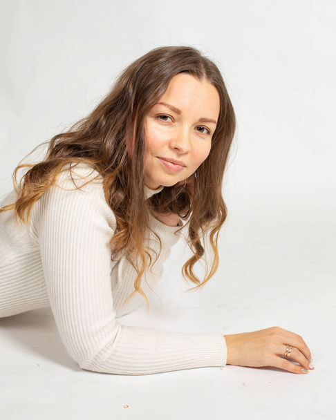 Porträt einer lächelnden attraktiven jungen Europäerin, die in einem warmen weißen Pullover auf weißem Grund liegt - Foto, Bild