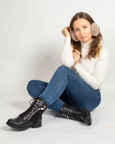 Young woman in warm white jumper with fur earmuffs isolated on white sitting on the floor in blue jeans - Foto, Imagem