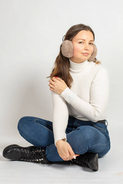 Young woman in warm white jumper with fur earmuffs isolated on white sitting on the floor with crossed legs in jeans - Foto, Imagem
