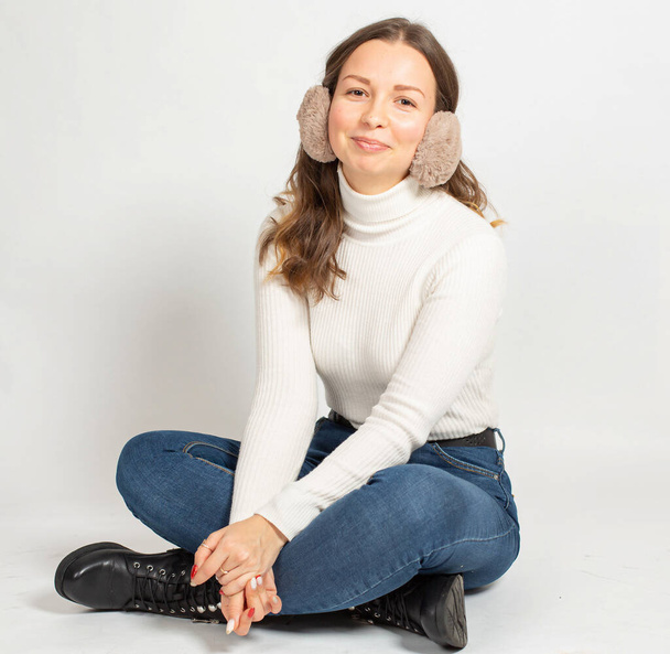 Smiling young woman in warm white jumper with fur earmuffs isolated on white sitting on the floor with crossed legs in jeans - Fotoğraf, Görsel